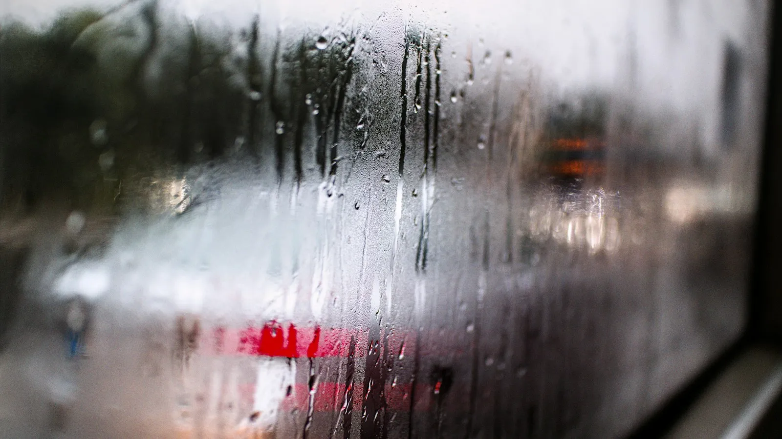 Rain and fog droplets streaking down a car window with blurred red lights behind it